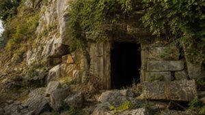 A realistic ancient stone cave threshold partly hidden by temple ruins and vegetation with no readable symbols.