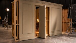 A realistic editorial image of a film set wall section with a concealed door being prepared under studio lights.
